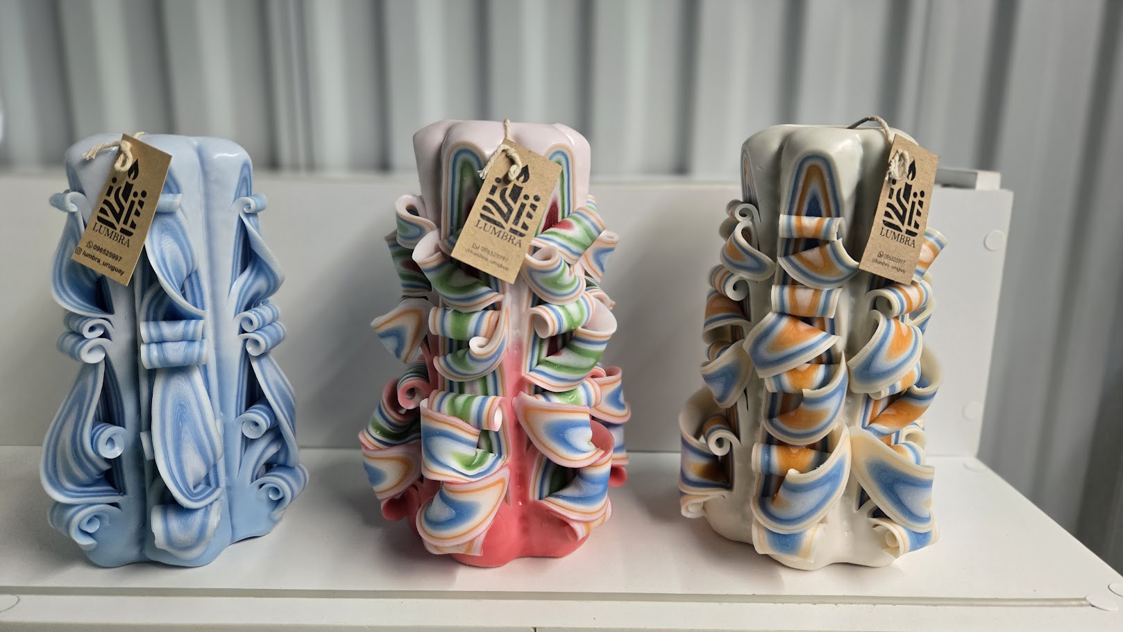 Tres velas artesanales talladas a mano de tamaño grande, con diseños decorativos diferentes en tonos celeste, multicolor y blanco con naranja, sobre fondo claro.